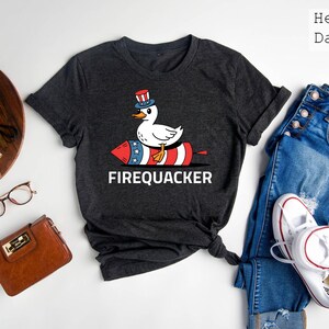 Peut inclure: T-shirt gris fonc&eacute; avec un canard de dessin anim&eacute; portant un chapeau patriotique et chevauchant un p&eacute;tard. Le mot "FIREQUACKER" est imprim&eacute; en blanc en dessous. Le t-shirt est associ&eacute; &agrave; un jean bleu et des baskets blanches.