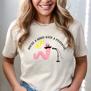 May include: A beige t-shirt with a pink worm with a black mustache and a microphone. The text "You're a worm with a mustache" is printed on the shirt.