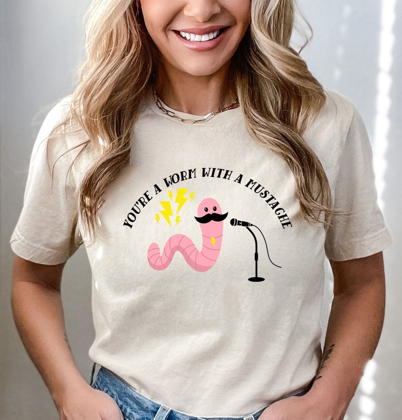 May include: A beige t-shirt with a pink worm with a black mustache and a microphone. The text "You're a worm with a mustache" is printed on the shirt.