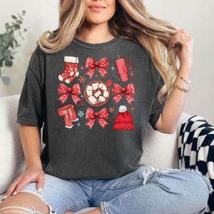 May include: Dark gray t-shirt featuring a festive graphic with red bows, a stocking, a scarf, a mug, and a beanie. The design includes a cup of hot chocolate with marshmallows and floral accents. The shirt is made of a soft material.