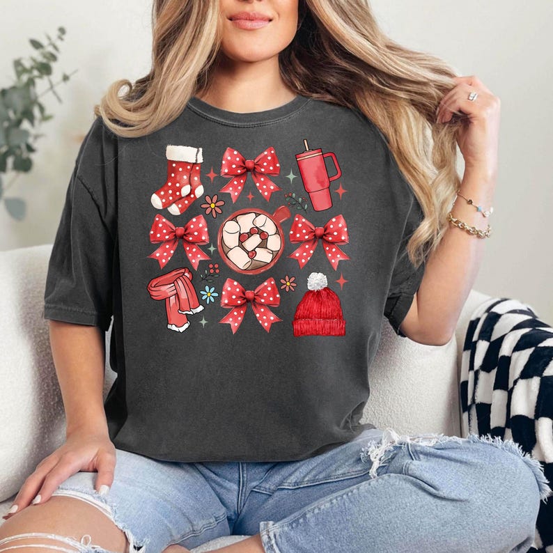 May include: Dark gray t-shirt featuring a festive graphic with red bows, a stocking, a scarf, a mug, and a beanie. The design includes a cup of hot chocolate with marshmallows and floral accents. The shirt is made of a soft material.