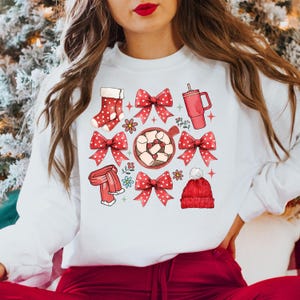 May include: White sweatshirt featuring a festive design with red bows, a stocking, a mug, a scarf, and a beanie. The design includes a cup of hot chocolate with marshmallows and floral accents, perfect for the holiday season.