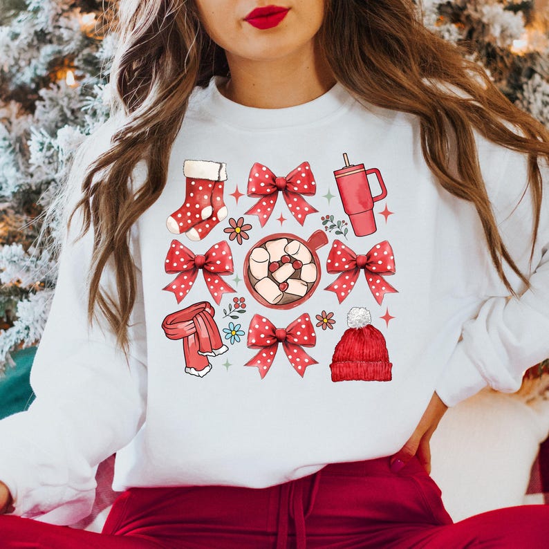 May include: White sweatshirt featuring a festive design with red bows, a stocking, a mug, a scarf, and a beanie. The design includes a cup of hot chocolate with marshmallows and floral accents, perfect for the holiday season.