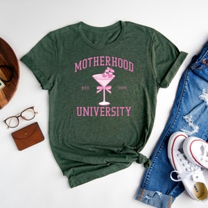 May include: A green t-shirt with the text "MOTHERHOOD UNIVERSITY" in pink lettering. The text is surrounded by a pink cocktail glass with a pink bow and two pink cherries. The text "EST. 1989" is below the cocktail glass.