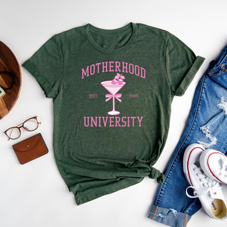 May include: A green t-shirt with the text "MOTHERHOOD UNIVERSITY" in pink lettering. The text is surrounded by a pink cocktail glass with a pink bow and two pink cherries. The text "EST. 1989" is below the cocktail glass.