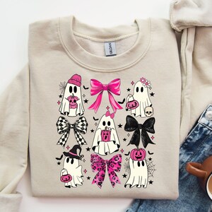 Puede incluir: Sudadera beige con un dise&ntilde;o tem&aacute;tico de Halloween. El dise&ntilde;o incluye fantasmas de dibujos animados con lazos, sombreros y calabazas. Los lazos son de varios estilos y colores, incluyendo rosa, negro y patrones a cuadros. Tambi&eacute;n se incluyen murci&eacute;lagos y estrellas negras.