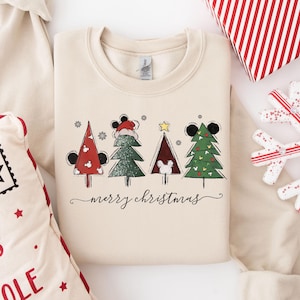 May include: A cream-coloured sweatshirt with a festive design. It features four Christmas trees adorned with Mickey Mouse ears and faces. The trees are red, green, and brown, with a gold star and snowflakes. The words "merry christmas" are written in a script font.