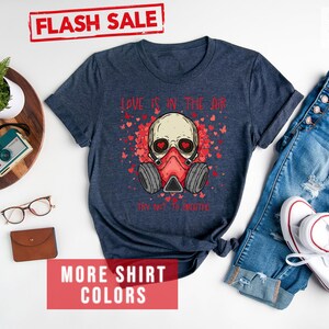 Pu&ograve; includere: Una t-shirt blu navy heather con un teschio che indossa una maschera antigas e il testo "Love is in the air. Try not to breathe." La t-shirt ha un design a cuore rosso e il testo &egrave; rosso. La t-shirt &egrave; su uno sfondo bianco con un paio di jeans, una macchina fotografica, un orologio e un paio di occhiali.