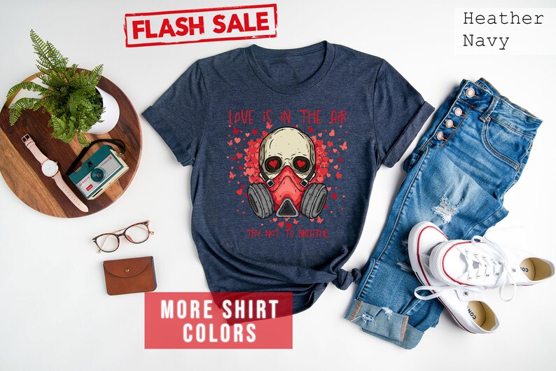 Pu&ograve; includere: Una t-shirt blu navy heather con un teschio che indossa una maschera antigas e il testo "Love is in the air. Try not to breathe." La t-shirt ha un design a cuore rosso e il testo &egrave; rosso. La t-shirt &egrave; su uno sfondo bianco con un paio di jeans, una macchina fotografica, un orologio e un paio di occhiali.