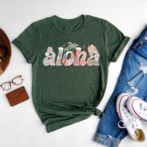 Puede incluir: Una camiseta verde con la palabra "Aloha" impresa en la parte delantera en un dise&ntilde;o floral. La palabra est&aacute; en una fuente rosa y blanca con hojas y flores verdes que la rodean.