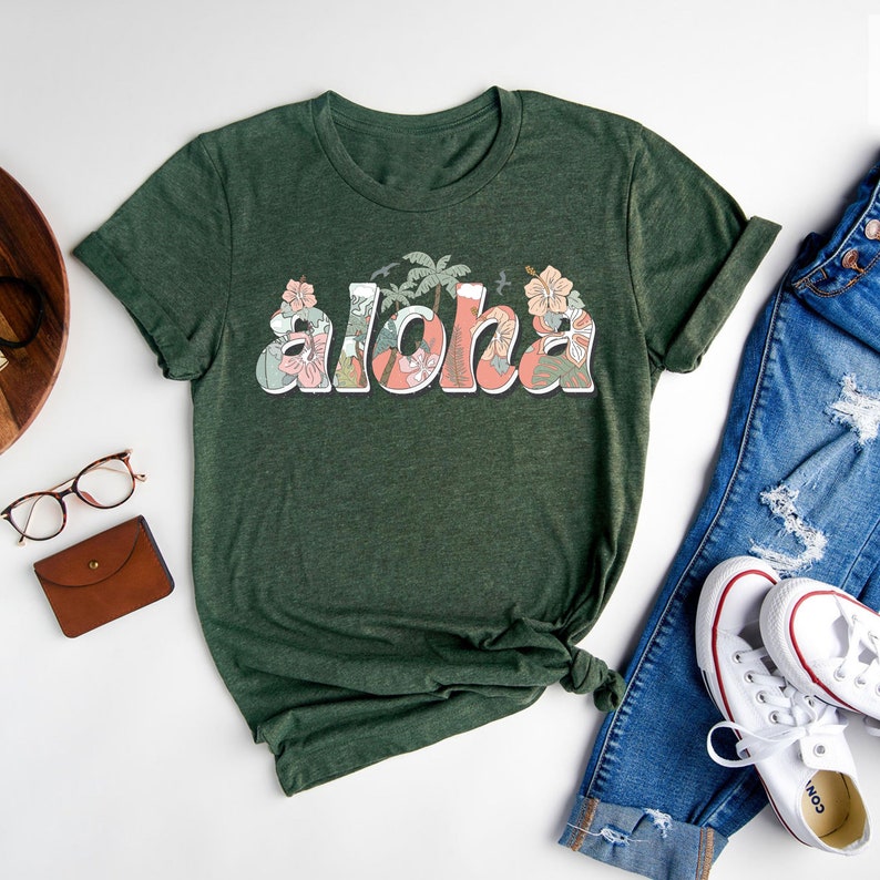 Puede incluir: Una camiseta verde con la palabra "Aloha" impresa en la parte delantera en un dise&ntilde;o floral. La palabra est&aacute; en una fuente rosa y blanca con hojas y flores verdes que la rodean.