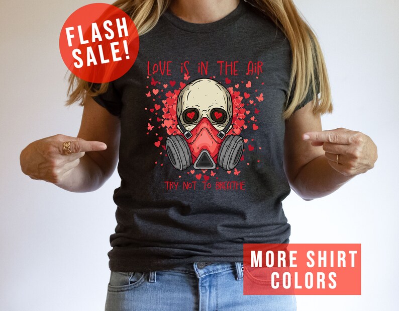 Pu&ograve; includere: Una maglietta grigia scura con una grafica di uno scheletro che indossa una maschera antigas e circondato da cuori rossi. Il testo "LOVE IS IN THE AIR" e "TRY NOT TO BREATHE" &egrave; stampato sulla maglietta.