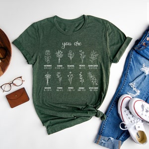 May include: A forest green t-shirt with white text that reads "you are" and features a list of words and corresponding bible verses about flowers. The shirt is laid out on a white surface with a pair of glasses, a brown wallet, a wooden tray with a camera and a plant, and a pair of white sneakers with blue jeans.