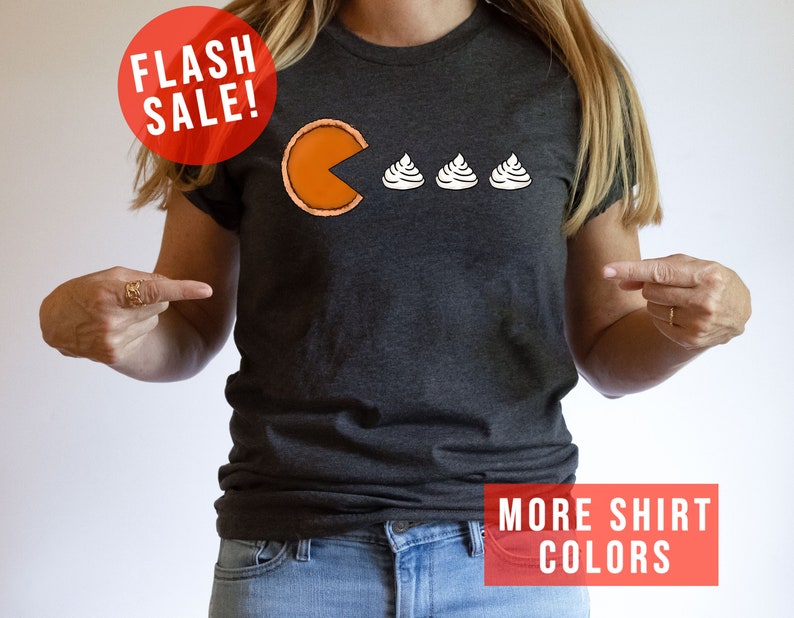 K&ouml;nnte beinhalten: Ein dunkelgraues T-Shirt mit einem Grafikdruck von einem Pac-Man, der drei Mini-K&uuml;rbiskuchen isst. Der Text "FLASH SALE!" befindet sich in einem roten Kreis oben im Bild. Der Text "MORE SHIRT COLORS" befindet sich in einem roten Rechteck unten im Bild.