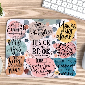 Op de afbeelding: Een kleurrijke muismat met inspirerende citaten en abstracte vormen. De citaten zijn "You are enough", "It's OK to not be OK", "Grow Positive Thoughts", en "Count your blessings, not your worries".