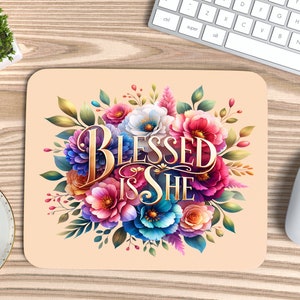 Op de afbeelding: Een roze en perzikkleurige muismat met een bloemenontwerp en de tekst "Blessed is She" in gouden letters.