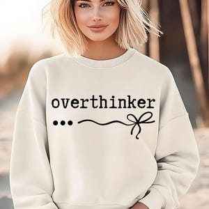 Peut inclure: Sweat-shirt blanc avec le texte "overthinker" en police d'écriture de machine à écrire noire, suivi de trois points noirs, une ligne ondulée et un nœud noir.