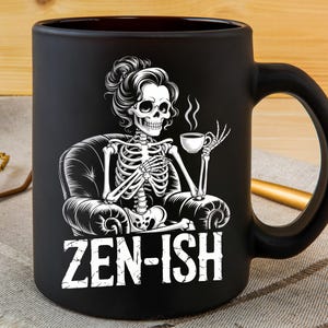 Op de afbeelding: Zwarte keramische mok met een witte illustratie van een skelet in een stoel, met een kopje koffie. De mok heeft de witte tekst "ZEN-ISH". De mok heeft een zwart handvat en staat op een houten oppervlak.