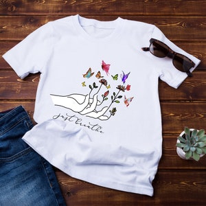 以下が含まれることがあります： 花と蝶々を持った手を描いた白いTシャツ。手の下に「Just Breathe」という文字がプリントされています。