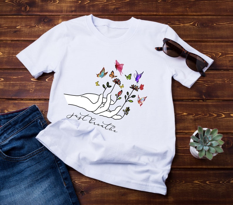 以下が含まれることがあります： 花と蝶々を持った手を描いた白いTシャツ。手の下に「Just Breathe」という文字がプリントされています。