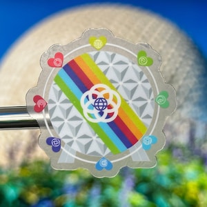 Könnte beinhalten: Ein silberner Aufkleber mit einem Regenbogen und einem weißen geometrischen Design. Der Aufkleber zeigt in jeder Ecke ein Herz mit einem Regenbogenmuster. Der Aufkleber befindet sich vor einem unscharfen Bild einer großen weißen Kugel.
