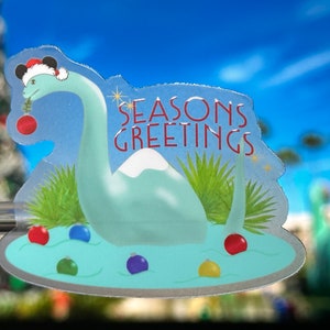 May include: A clear plastic holiday decoration featuring a light blue dinosaur wearing a Santa hat with Mickey Mouse ears. The dinosaur is in a lake with colorful ornaments. The text "SEASONS GREETINGS" is above the dinosaur.