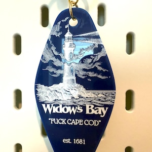 May include: A dark blue, diamond-shaped motel-style keychain featuring a white engraved illustration of a lighthouse on a rocky shore. The text reads "Widow's Bay", "FUCK CAPE COD", and "est. 1681" in white lettering. A gold-colored metal ring is attached.