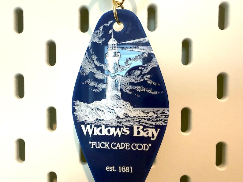 May include: A dark blue, diamond-shaped motel-style keychain featuring a white engraved illustration of a lighthouse on a rocky shore. The text reads "Widow's Bay", "FUCK CAPE COD", and "est. 1681" in white lettering. A gold-colored metal ring is attached.