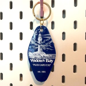 May include: A blue, diamond-shaped motel-style keychain featuring a white graphic of a lighthouse on a rocky shore. The text reads "Widow's Bay", "FUCK CAPE COD", and "est. 1681" in white lettering. It includes a gold-toned metal split ring for keys.