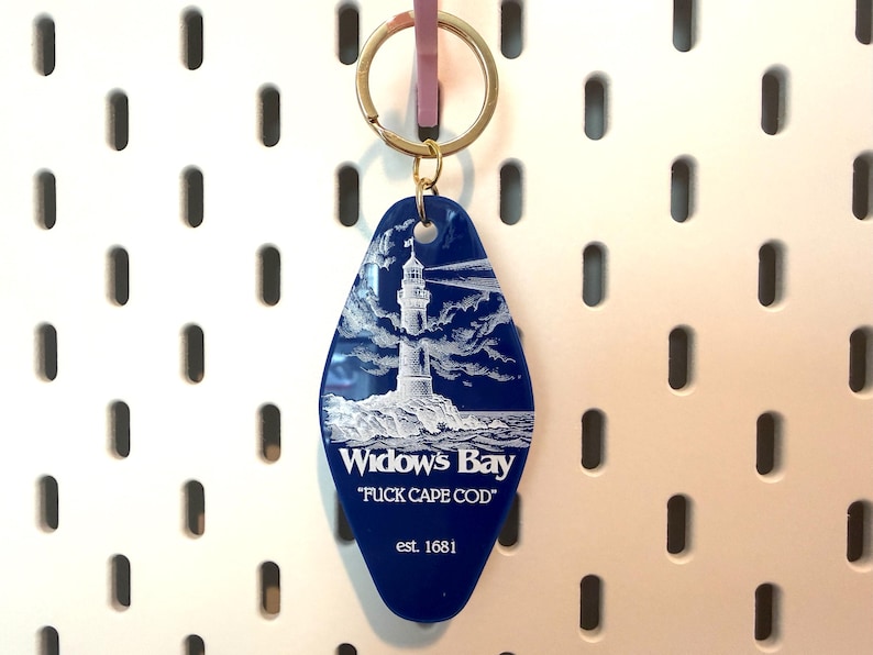 May include: A blue, diamond-shaped motel-style keychain featuring a white graphic of a lighthouse on a rocky shore. The text reads "Widow's Bay", "FUCK CAPE COD", and "est. 1681" in white lettering. It includes a gold-toned metal split ring for keys.