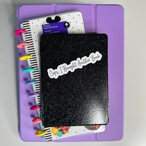 May include: A stack of stationery items, including a black glitter-covered notebook with the text "Oops, I Bought Another Book" on a white sticker. A purple tablet case and a planner with colorful disc binding are also visible.
