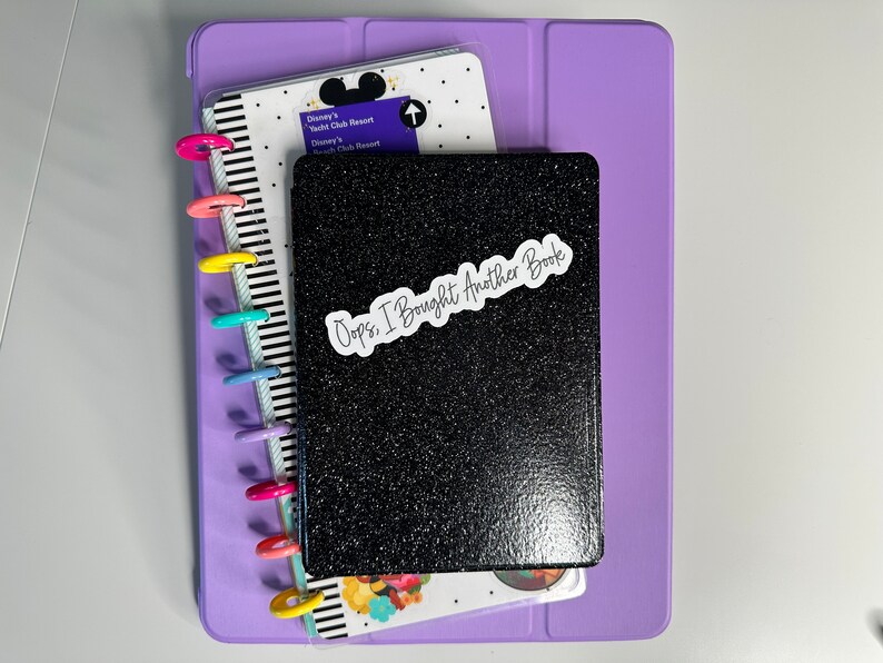 May include: A stack of stationery items, including a black glitter-covered notebook with the text "Oops, I Bought Another Book" on a white sticker. A purple tablet case and a planner with colorful disc binding are also visible.