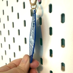 May include: A blue acrylic keychain with a white printed design of a tree and text. The keychain is diamond-shaped and attached to a gold-colored metal jump ring and lobster clasp. The design features a detailed tree illustration and white text.