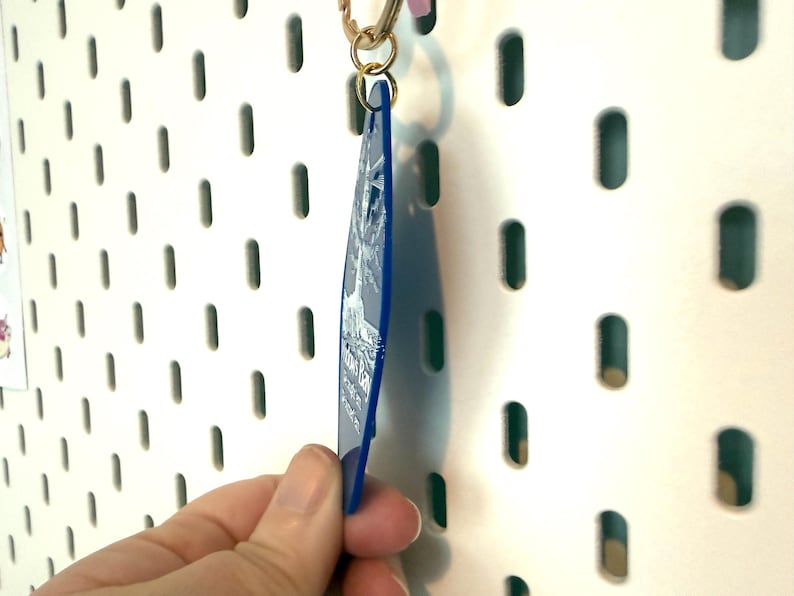 May include: A blue acrylic keychain with a white printed design of a tree and text. The keychain is diamond-shaped and attached to a gold-colored metal jump ring and lobster clasp. The design features a detailed tree illustration and white text.