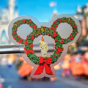 May include: A silver Mickey Mouse-shaped ornament with a red bow and a white candle in the center. The ornament is decorated with a green wreath and red poinsettia flowers.