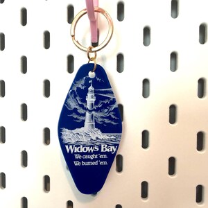 May include: A blue motel-style keychain with a gold-toned metal ring. The keychain features a white engraved illustration of a lighthouse on a rocky shore and the text: Widow's Bay, We caught 'em, We burned 'em.