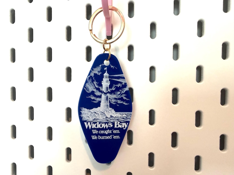 May include: A blue motel-style keychain with a gold-toned metal ring. The keychain features a white engraved illustration of a lighthouse on a rocky shore and the text: Widow's Bay, We caught 'em, We burned 'em.