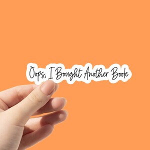 May include: A white sticker with the text "Oops, I Bought Another Book" in a handwritten style, held against an orange background. The sticker has a wavy outline.