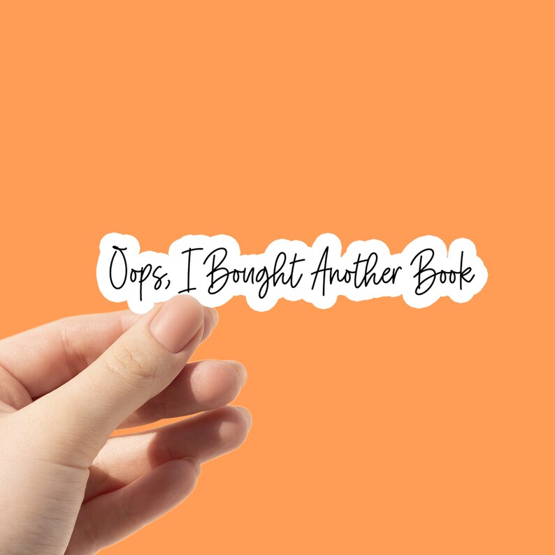 May include: A white sticker with the text "Oops, I Bought Another Book" in a handwritten style, held against an orange background. The sticker has a wavy outline.