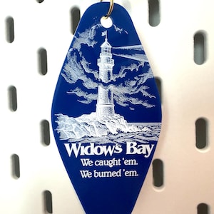 May include: A diamond-shaped blue motel-style keychain featuring a white graphic of a lighthouse on a rocky shore. The text reads Widow's Bay, We caught 'em. We burned 'em. A gold-colored metal ring is attached to the top of the blue plastic tag.