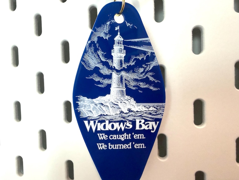 May include: A diamond-shaped blue motel-style keychain featuring a white graphic of a lighthouse on a rocky shore. The text reads Widow's Bay, We caught 'em. We burned 'em. A gold-colored metal ring is attached to the top of the blue plastic tag.