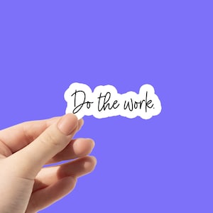 May include: A hand holds up a white sticker with the text "Do the work." on a purple background.