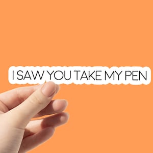May include: White sticker with black text that reads "I saw you take my pen". A hand is holding the sticker.