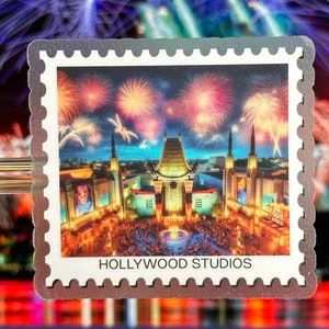 May include: A vintage-style postage stamp featuring a nighttime view of the Hollywood Hills with fireworks exploding in the sky. The text "HOLLYWOOD STUDIOS" is printed at the bottom of the stamp.