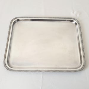 Puede incluir: Una bandeja rectangular plateada con un borde decorativo. La bandeja tiene esquinas redondeadas y una superficie lisa y reflectante. El borde presenta un patrón geométrico repetitivo.