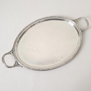 Peut inclure: Plateau de service ovale en argent avec deux poignées ornées. Le plateau a une surface réfléchissante et une bordure décorative avec un motif répétitif. Les poignées sont bouclées et détaillées, ajoutant au design élégant du plateau.