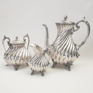 Op de afbeelding: Een zilveren theeservies met een theepot, een melkkan en een suikerpot. De set heeft een gegroefd ontwerp en sierlijke handvatten. De theepot heeft een lange tuit en een deksel met een knop.
