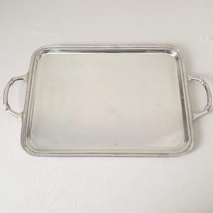 Könnte beinhalten: Ein rechteckiges silbernes Serviertablett mit zwei gebogenen Griffen. Das Tablett hat eine reflektierende Oberfläche und einen erhöhten Rand. Die Griffe sind an den Seiten des Tabletts befestigt und haben ein dekoratives Design.