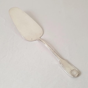 Pode incluir: Servidor de bolo prateado com uma lâmina larga e plana e um cabo longo e fino. A alça apresenta um design decorativo de concha na extremidade. O servidor é feito de um metal brilhante e reflexivo.
