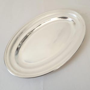 Könnte beinhalten: Eine silberfarbene, ovale Servierplatte. Die Platte hat eine glatte, reflektierende Oberfläche mit einem leicht erhöhten Rand und einem dezenten, geriffelten Design am Rand. Die Form ist länglich.